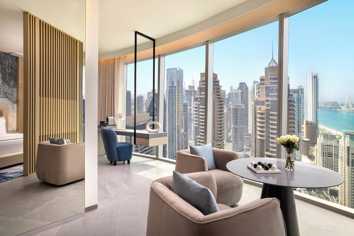 A modern hotel room with a large window offering a city view, a round table with a vase of flowers, and a sofa with cushions.