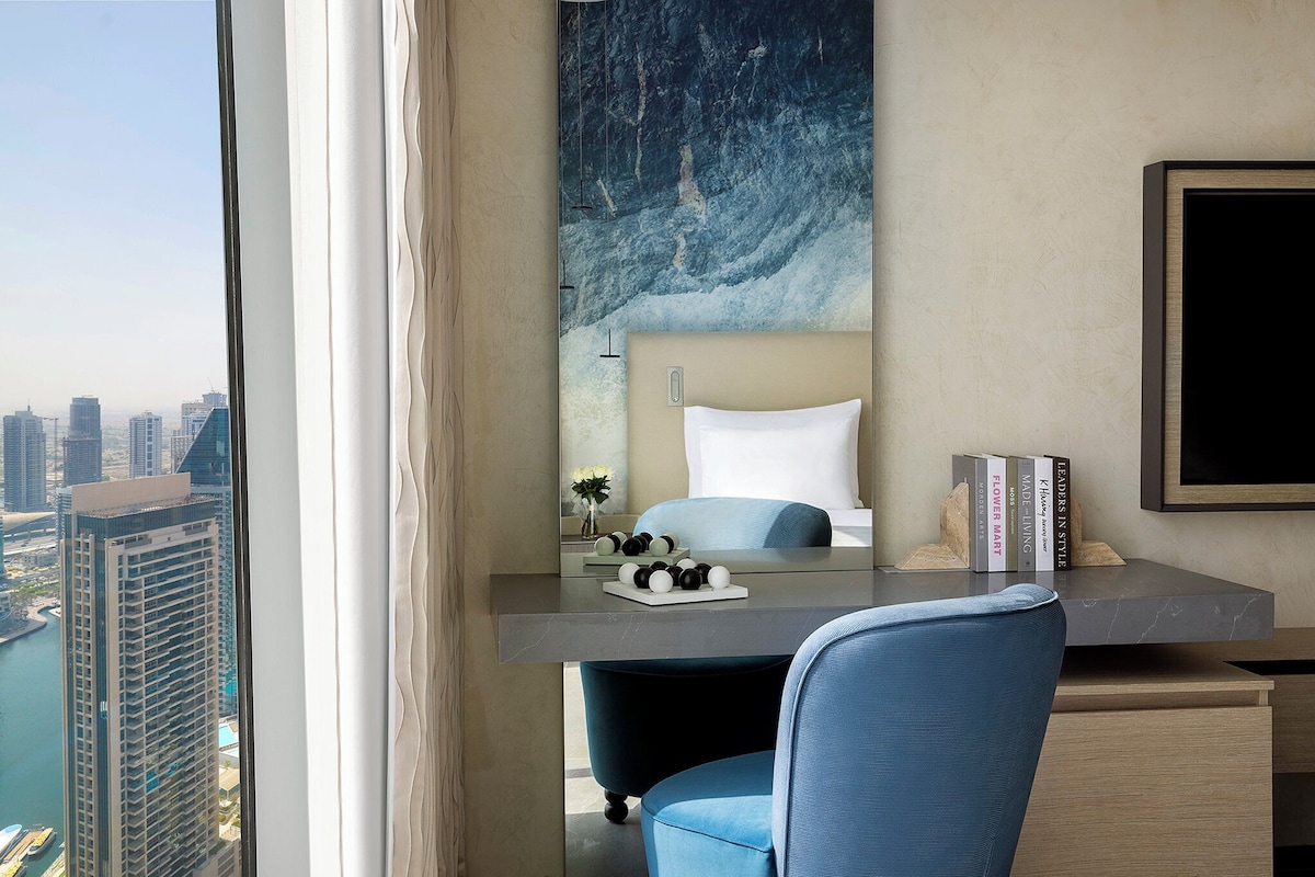 Ein modernes Hotelzimmer mit Blick auf die Stadt, ein Schreibtisch mit einem Laptop, ein Sessel, ein Bett und ein großes abstraktes Gemälde an der Wand.