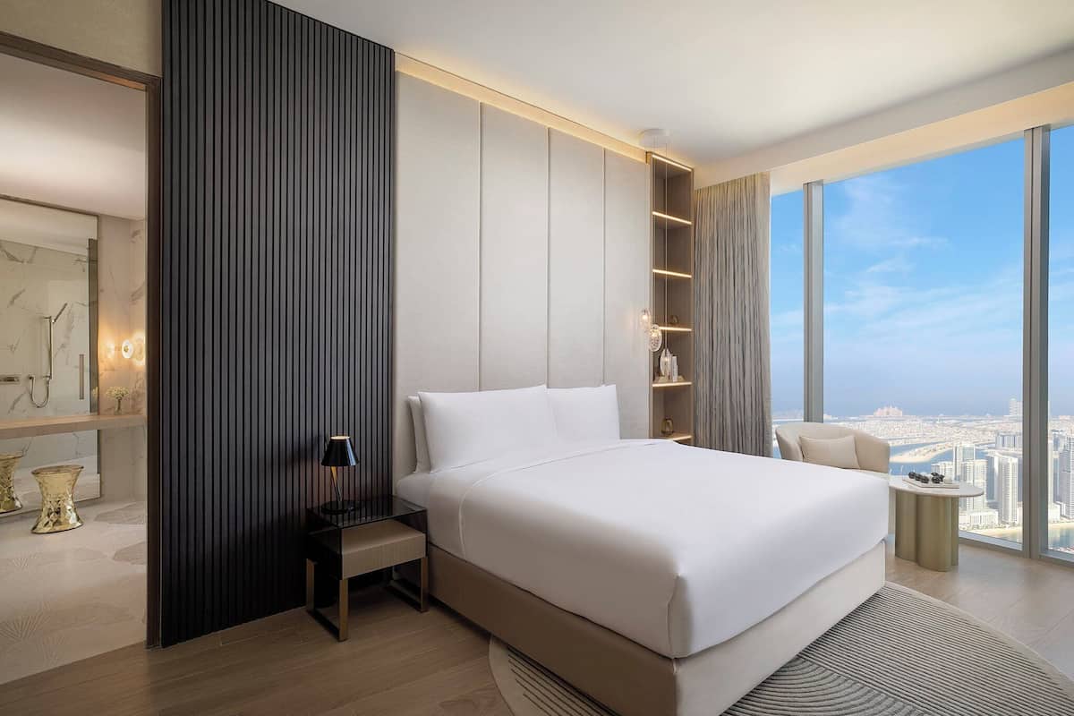 A modern hotel room with a large bed, a nightstand, a small table, and a view of the city through large windows.