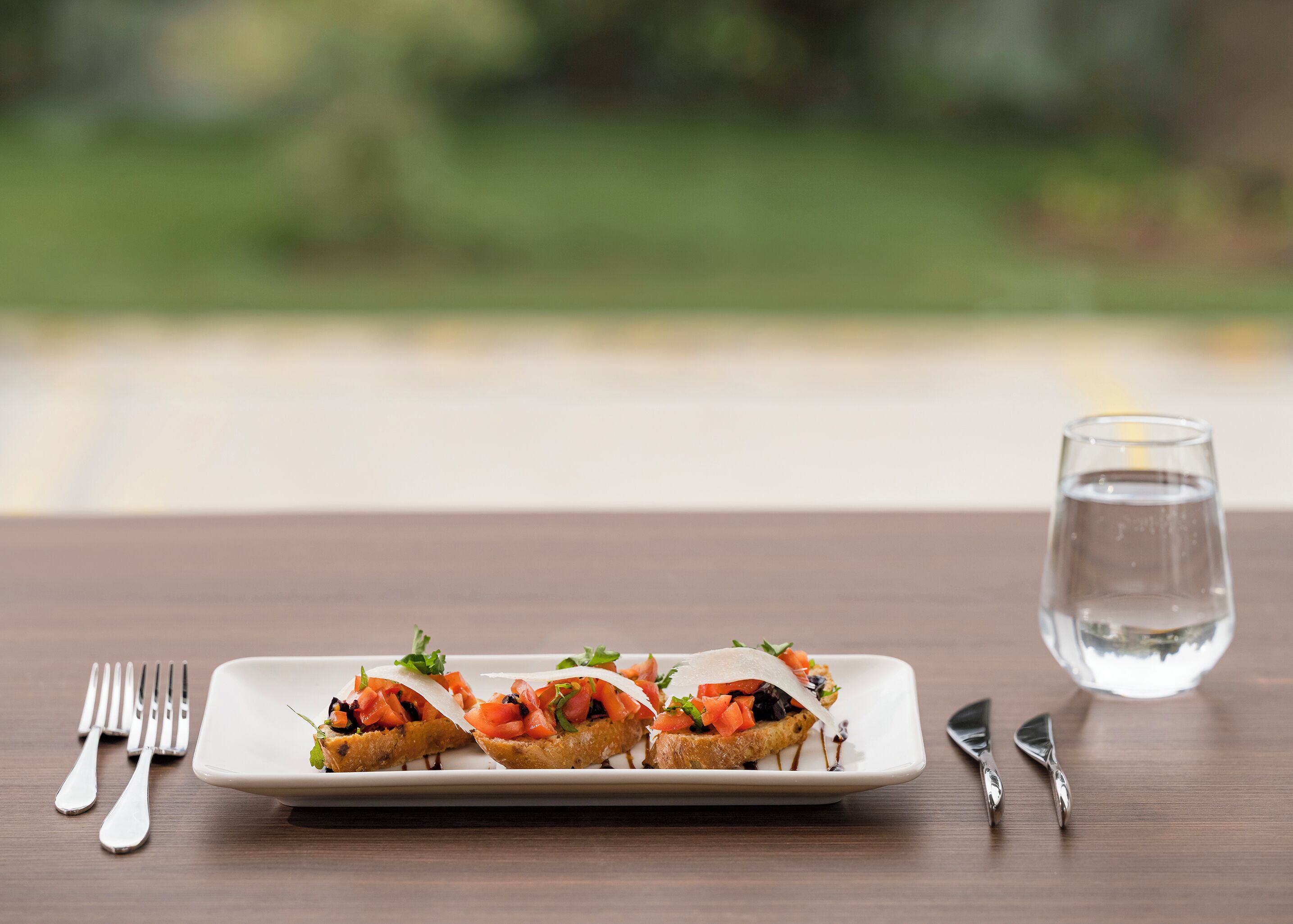 A rectangular white plate with two open-faced sandwiches garnished with herbs, accompanied by a glass of water and cutlery on a wooden table.