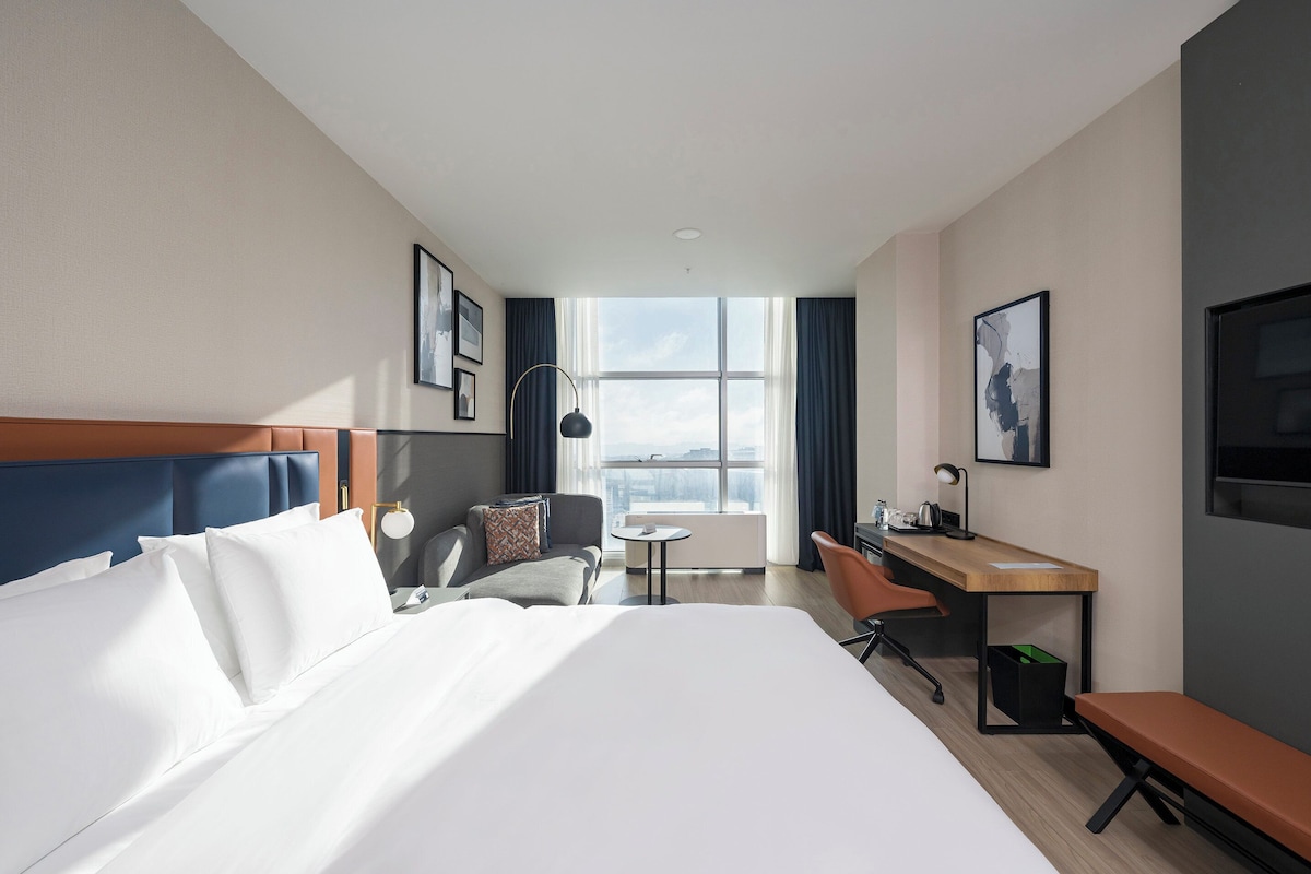Une chambre d’hôtel avec un grand lit, un bureau, une chaise, une petite table et une vue sur la ville.