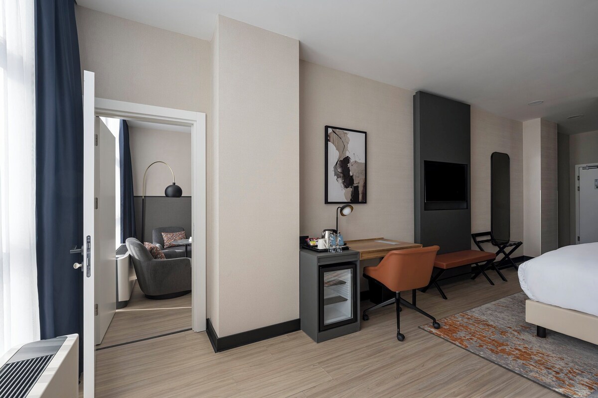 Habitación de hotel con cama, escritorio, silla, televisor, un refrigerador pequeño y una puerta que da a otra habitación.