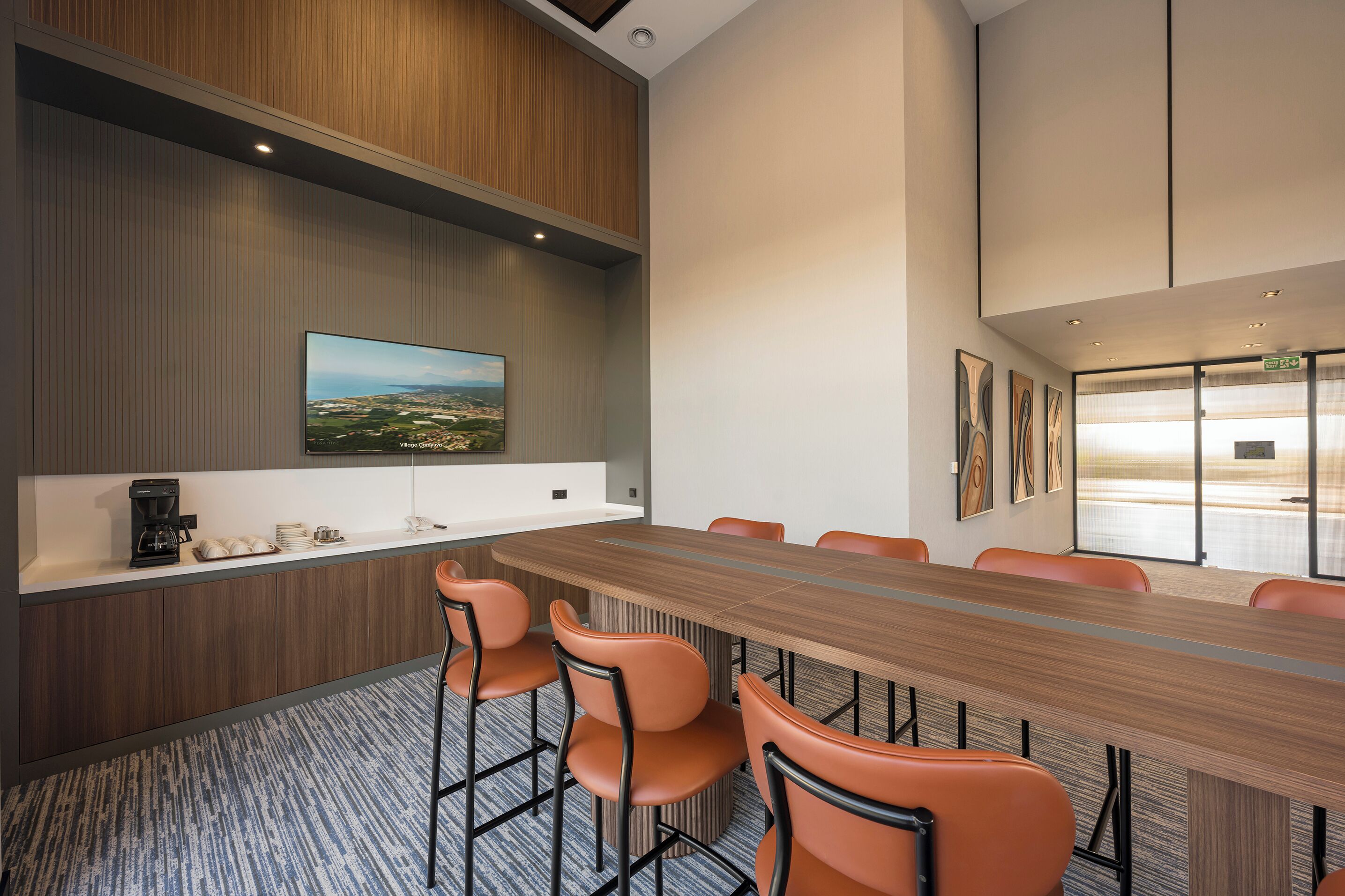 A conference room with a long table, six chairs, a coffee machine, and a mounted TV displaying a landscape.