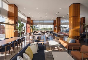 Een ruime binnenruimte met hoge plafonds, grote ramen en diverse zithoeken, waaronder tafels, stoelen en banken.