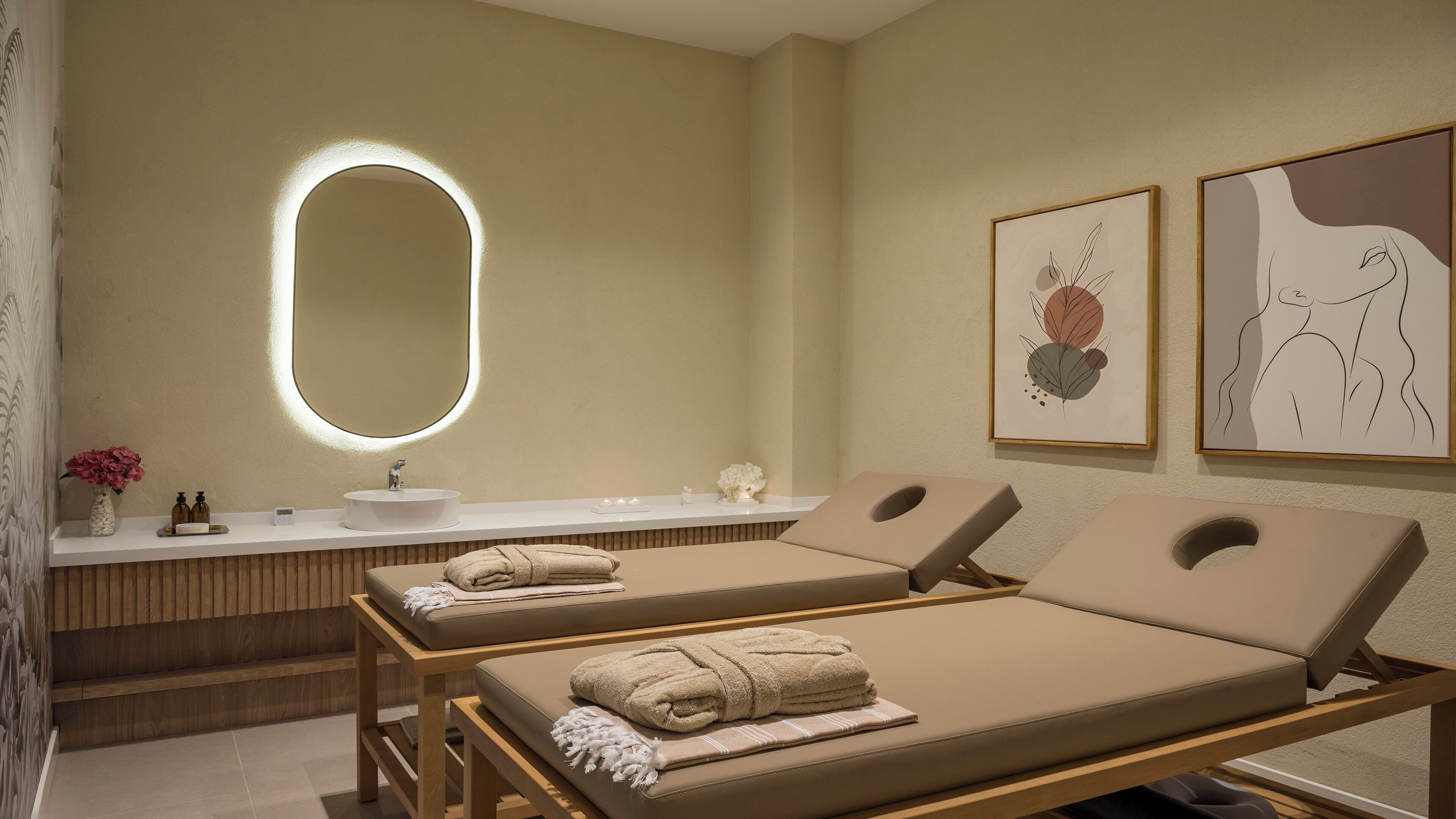 A spa room with a large mirror, two massage tables, a wooden bench, and artwork on the wall.
