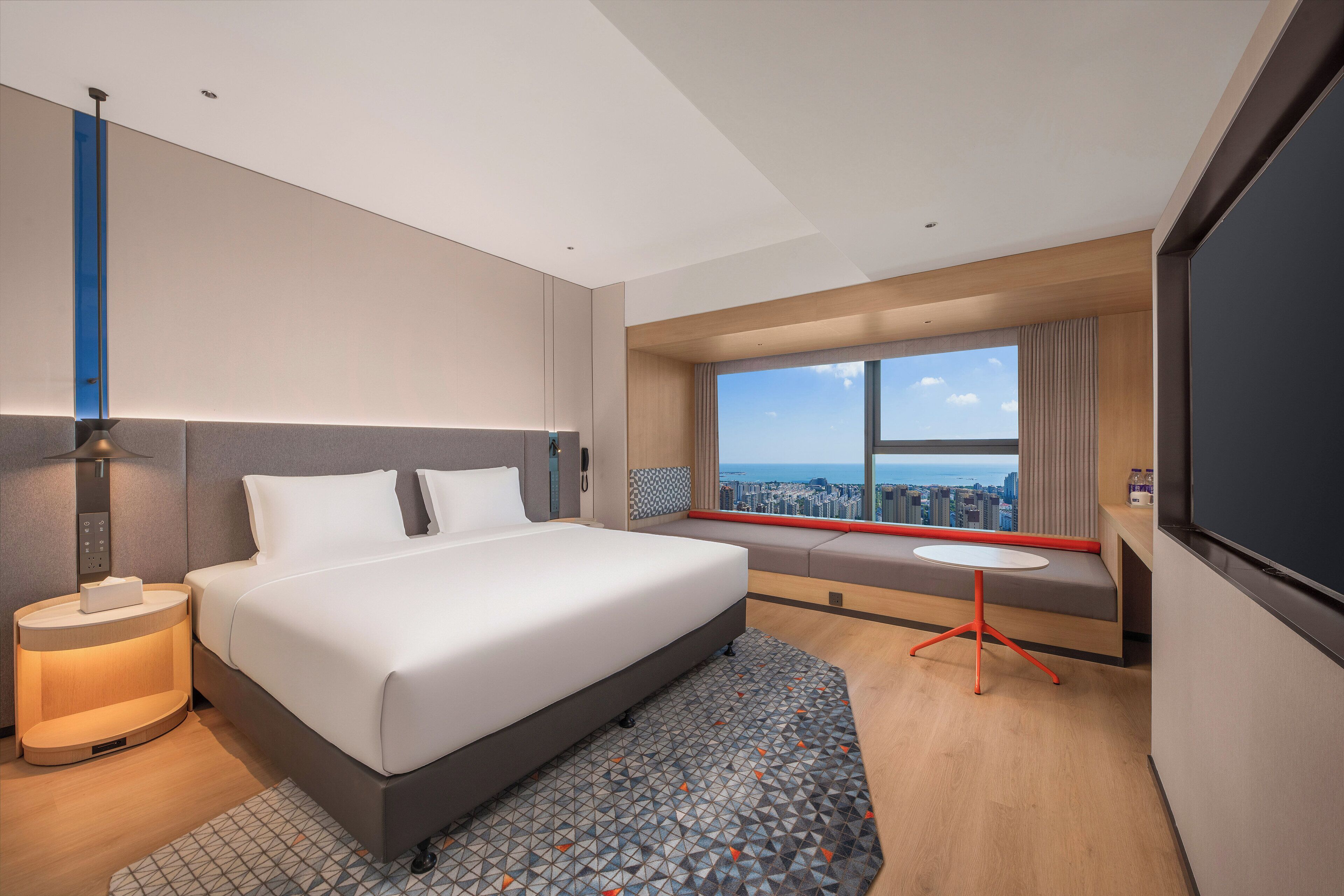A modern hotel room with a large bed, bedside table, and a view of the city.