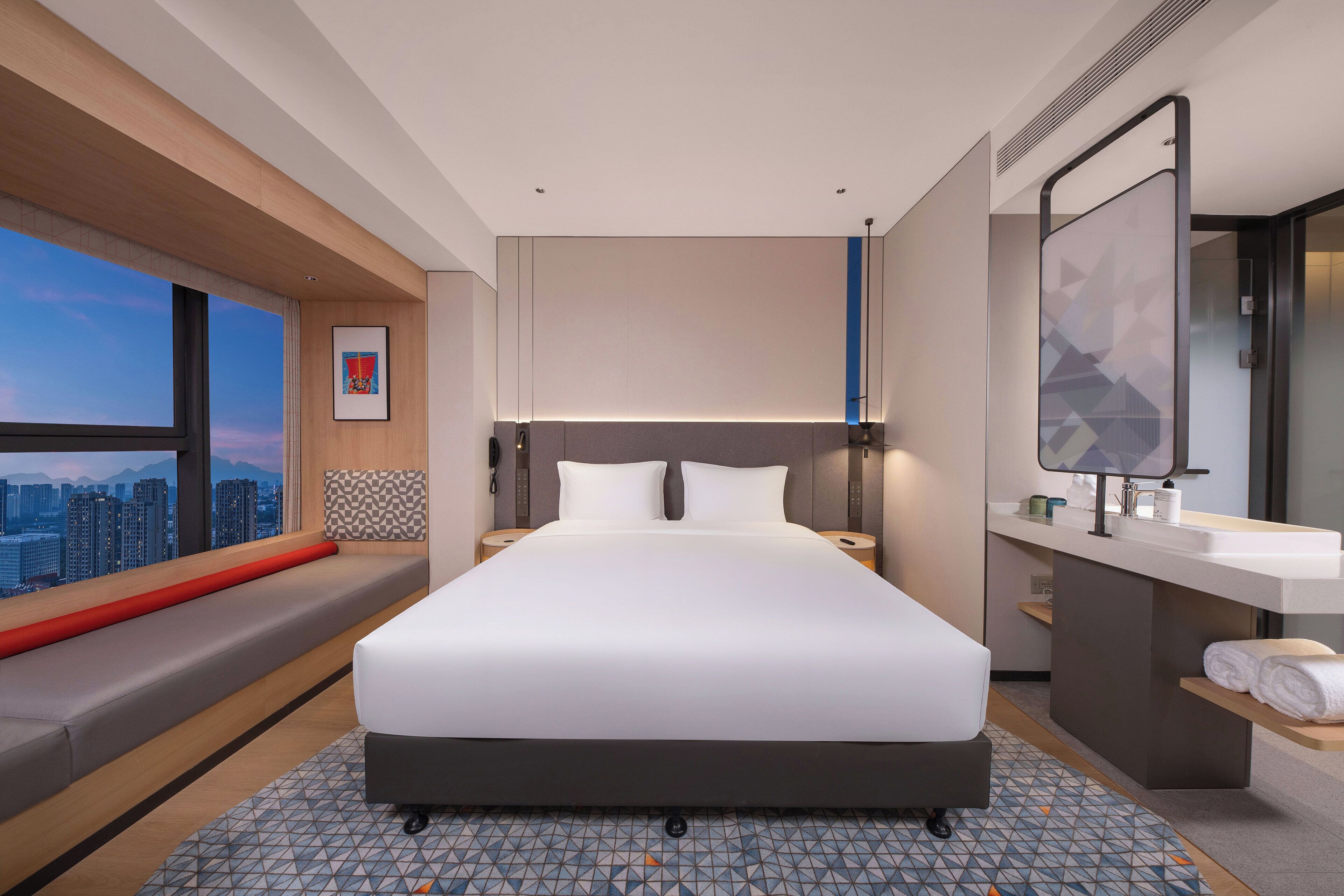 A modern hotel room with a large bed, a built-in bench along the window, a bathroom with a large mirror and sink, and a city view.