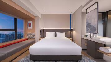 A modern hotel room with a large bed, a built-in bench along the window, a bathroom with a large mirror and sink, and a city view.