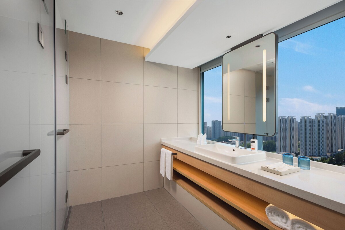 A modern bathroom with a large mirror, a white sink, and a view of tall buildings.
