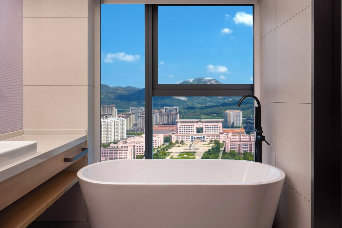 A modern bathroom with a freestanding bathtub, a large window offering a cityscape view, and a minimalist design.