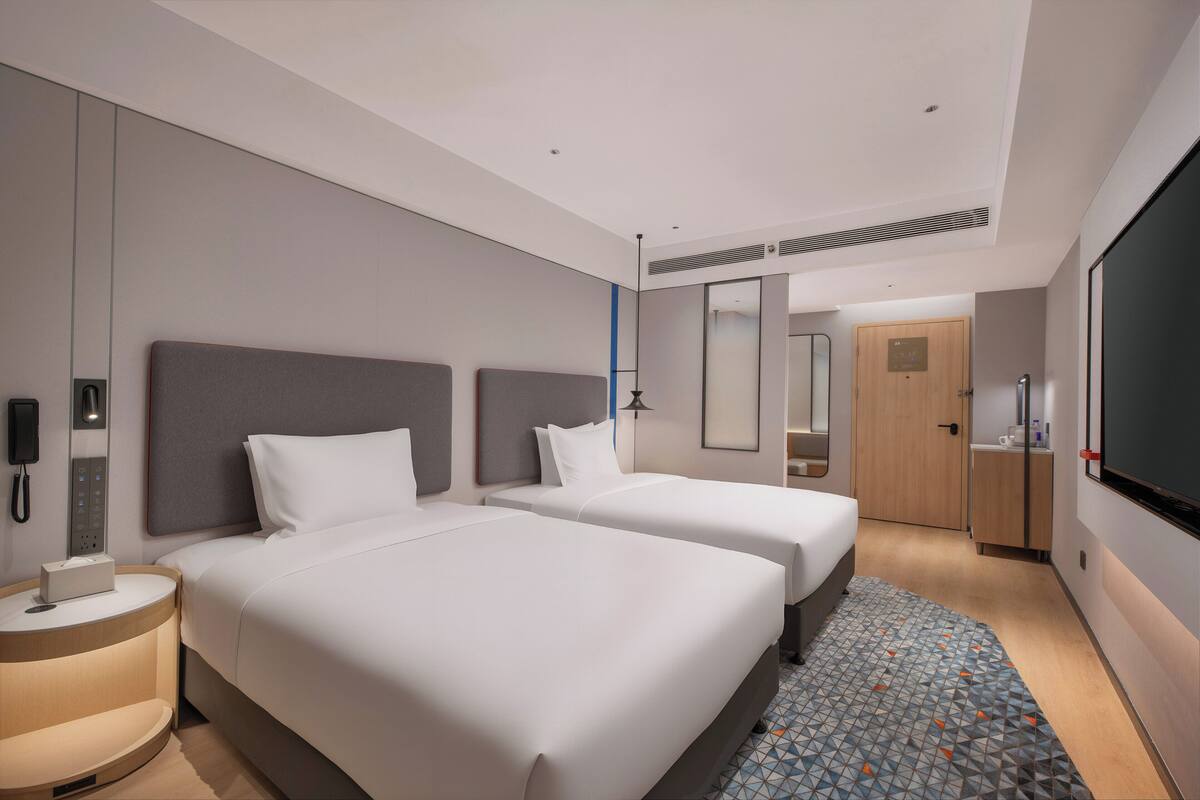 Ein Hotelzimmer mit zwei Betten, einem Nachttisch, einer Lampe, einem Kleiderschrank, einem Spiegel und einem Fernseher.