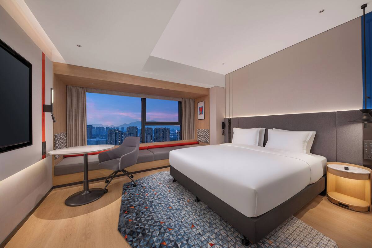 Ein Hotelzimmer mit einem großen Bett, einem Schreibtisch, einem Stuhl, einem kleinen Tisch, einem Nachttisch und Blick auf die Stadt.