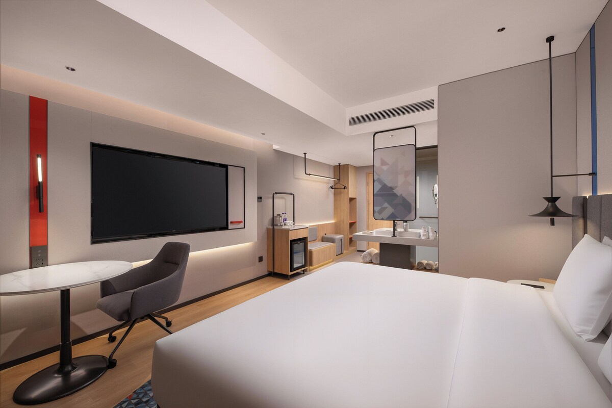 A modern hotel room with a large bed, a desk with a chair, a flat-screen TV, a small kitchenette, and a bathroom with a mirror and sink.