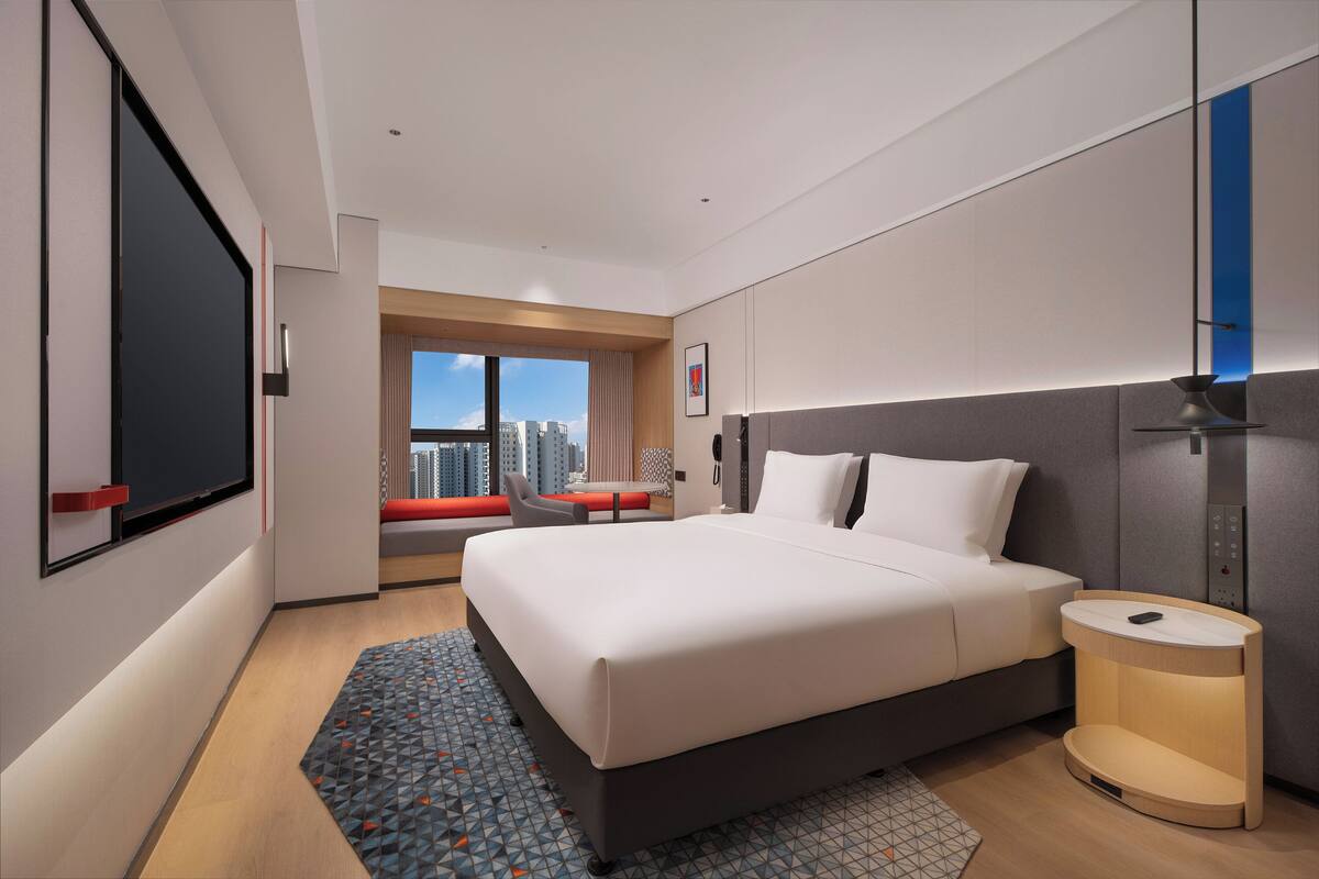 Ein modernes Hotelzimmer mit einem großen Bett, einem Nachttisch, einem Fenster mit Blick auf die Stadt und einem roten Sessel am Fenster.