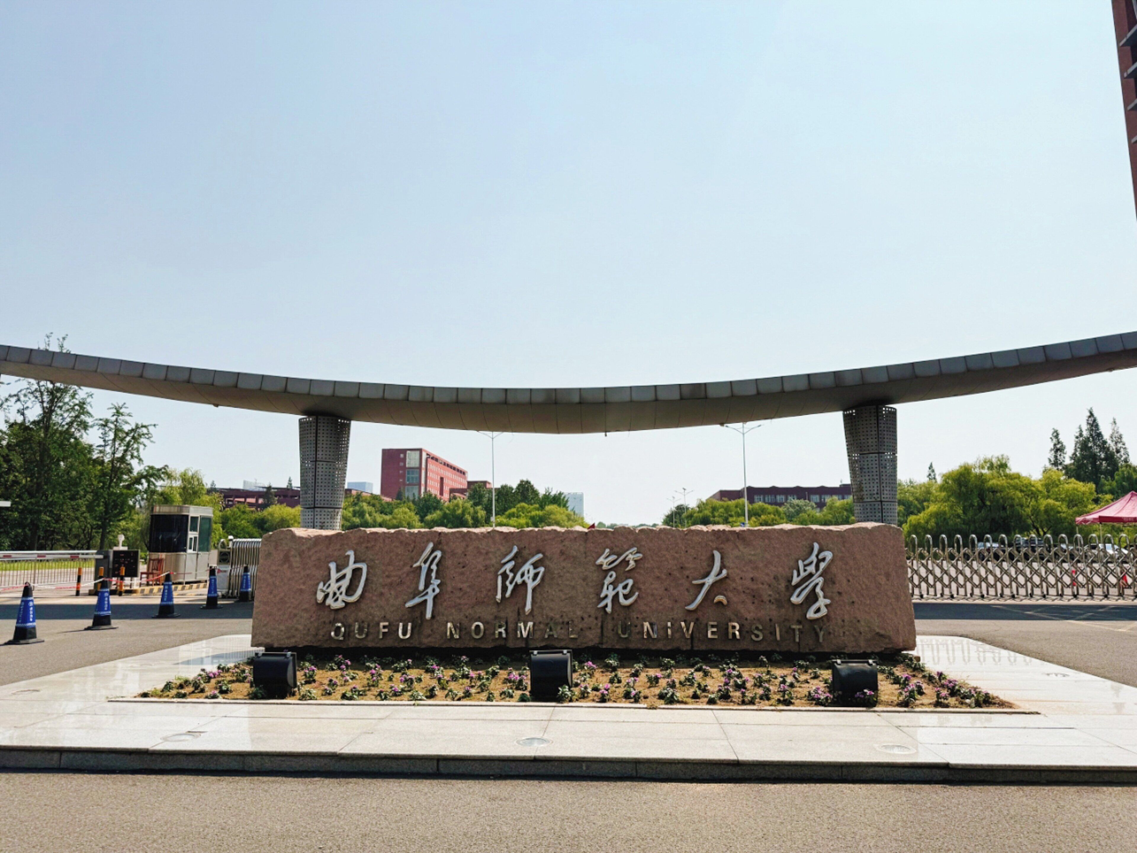 A stone sign with "QUFU NORMAN UNIVERSITY" written on it, surrounded by a landscaped area with flowers and a curved structure above.