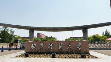 A stone sign with "QUFU NORMAN UNIVERSITY" written on it, surrounded by a landscaped area with flowers and a curved structure above.