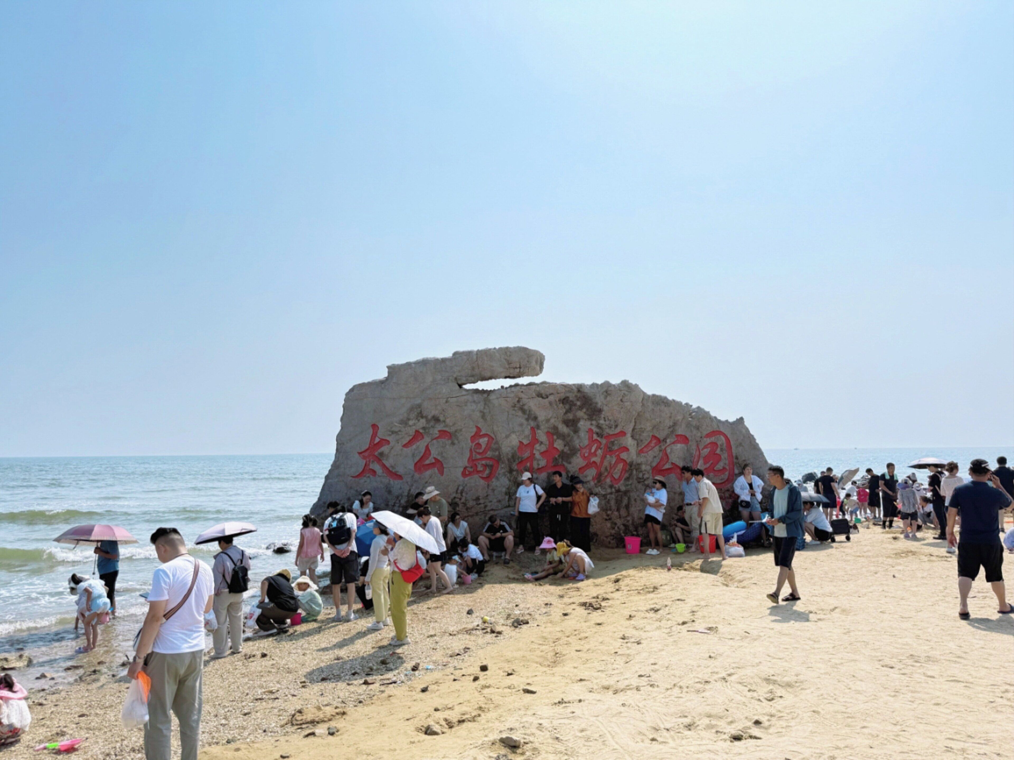 A beach with a large rock formation and people gathered around it.
