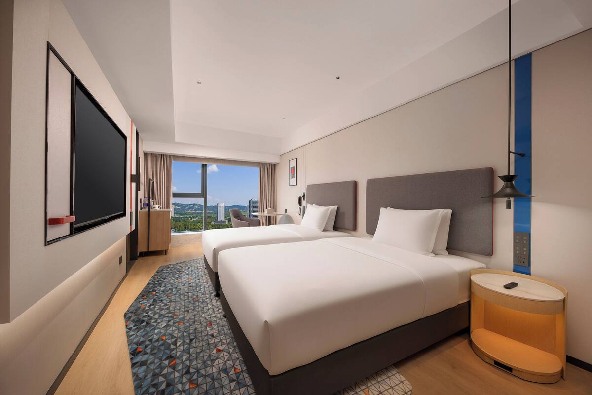 Ein Hotelzimmer mit zwei Betten, einem großen Fenster, einem Schreibtisch und Blick auf die Stadt.