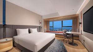A modern hotel room with a large bed, a desk, a chair, a TV, and a view of the city.