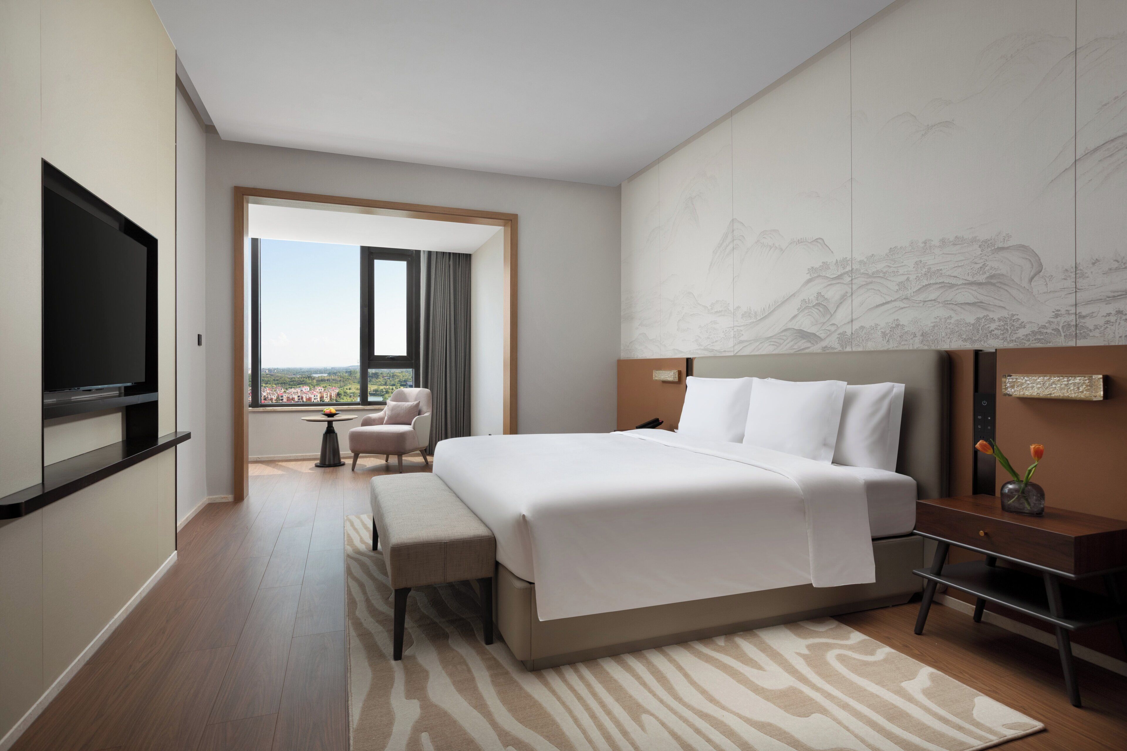 Une chambre d’hôtel avec un grand lit, une télévision, un bureau agrémenté d’un vase, et une vue sur le paysage urbain par la fenêtre.