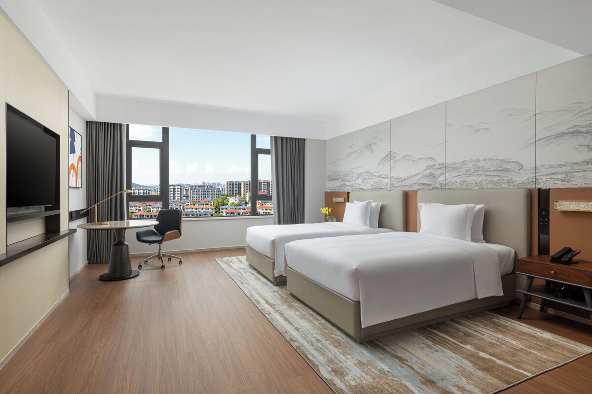 Ein Hotelzimmer mit zwei Betten, einem Schreibtisch, einem Sessel, einem Fernseher und einem großen Fenster mit Blick auf die Stadt.