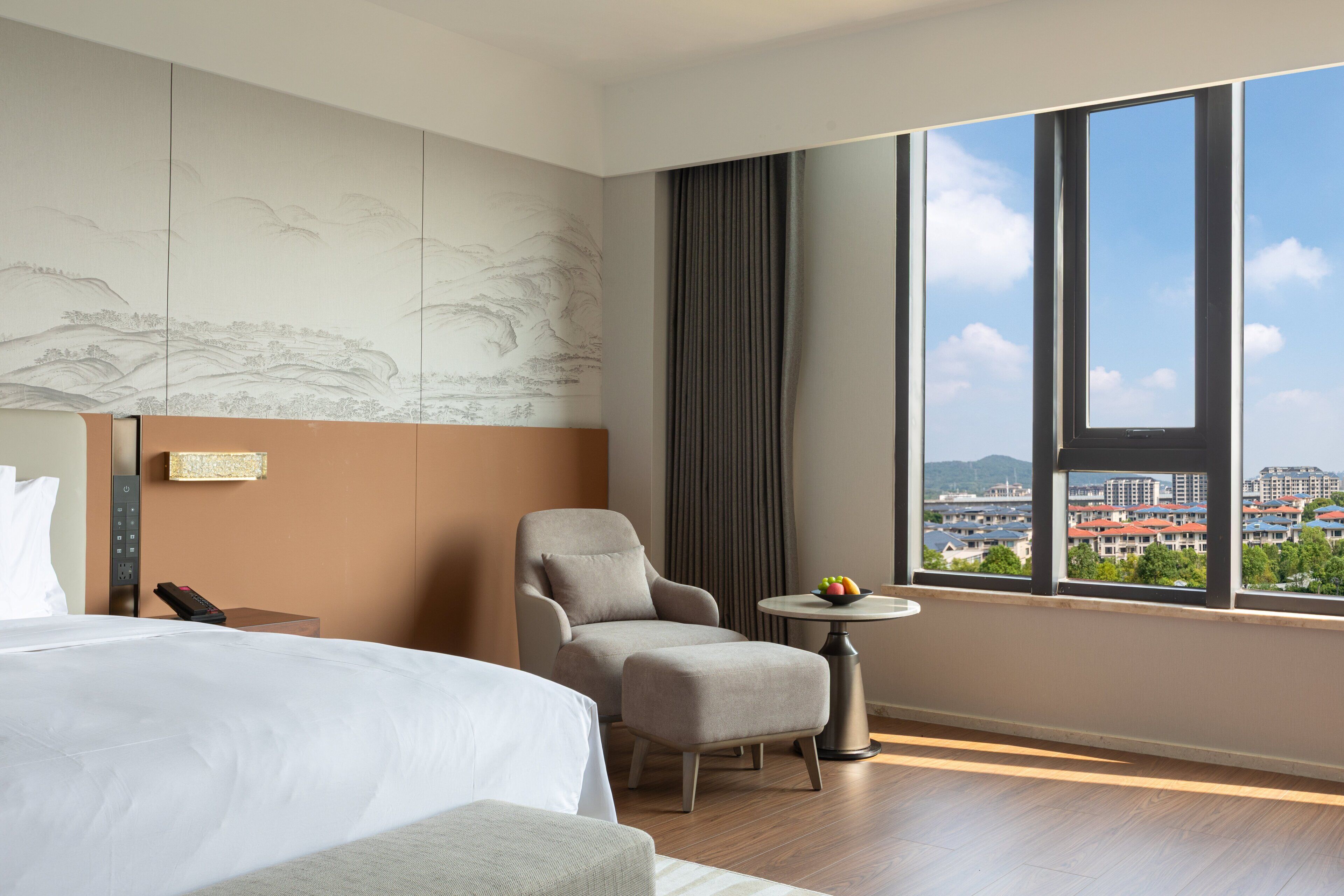 A hotel room with a bed, a chair, a small table with fruit, a view of buildings, and a wall with a landscape mural.