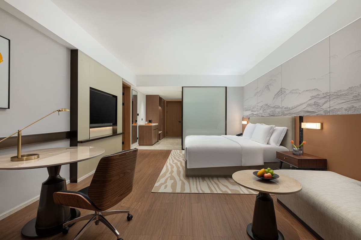 Une chambre d’hôtel avec un lit, un bureau, une chaise, une petite table avec des fruits et un mur orné d’un tableau représentant un paysage.