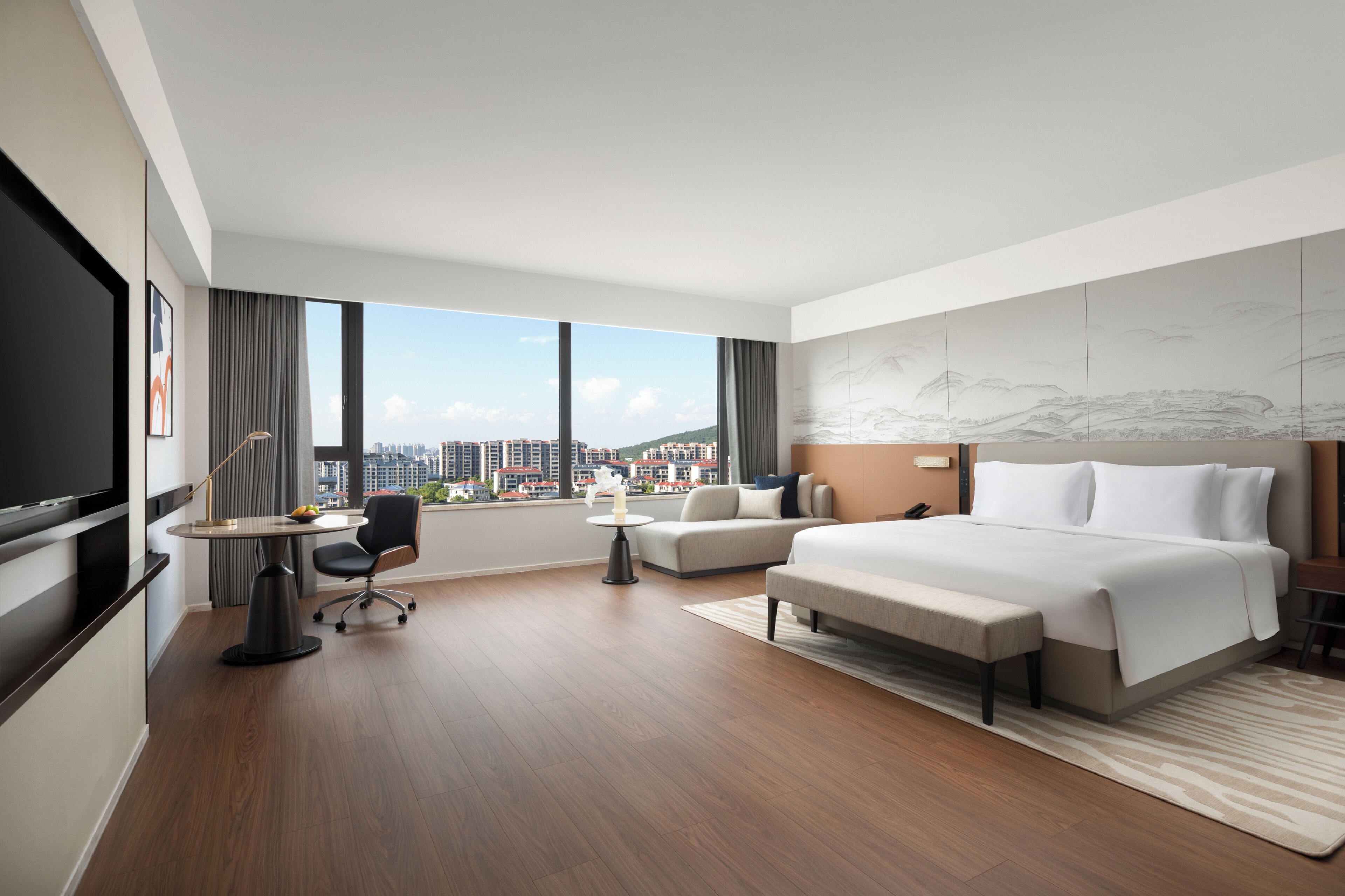 A modern hotel room with a large bed, a desk, a chair, a TV, and a view of the city.