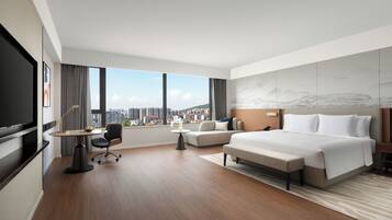 A modern hotel room with a large bed, a desk, a chair, a TV, and a view of the city.