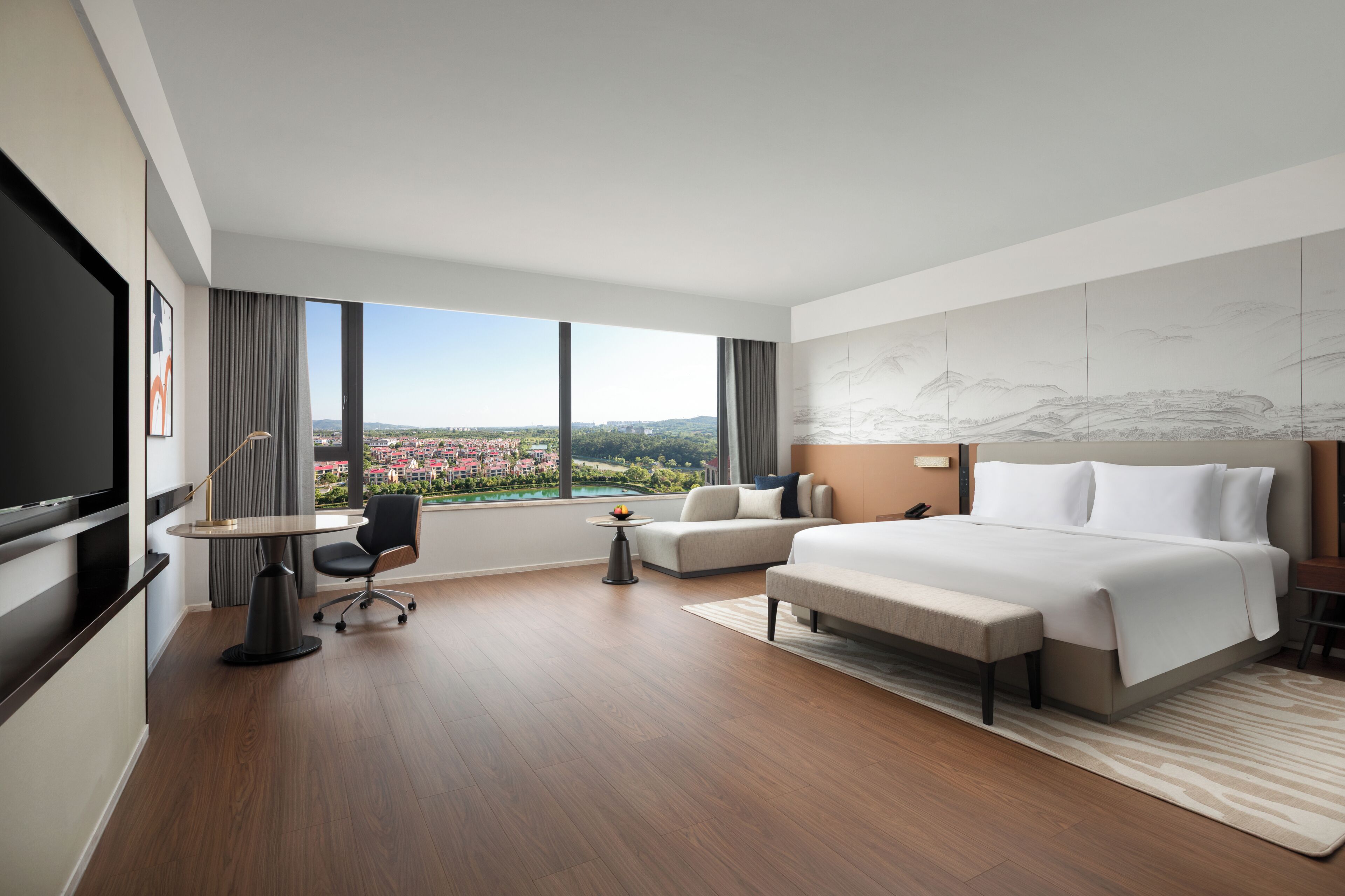 A modern hotel room with a large bed, a desk, a chair, a TV, and a view of a cityscape.