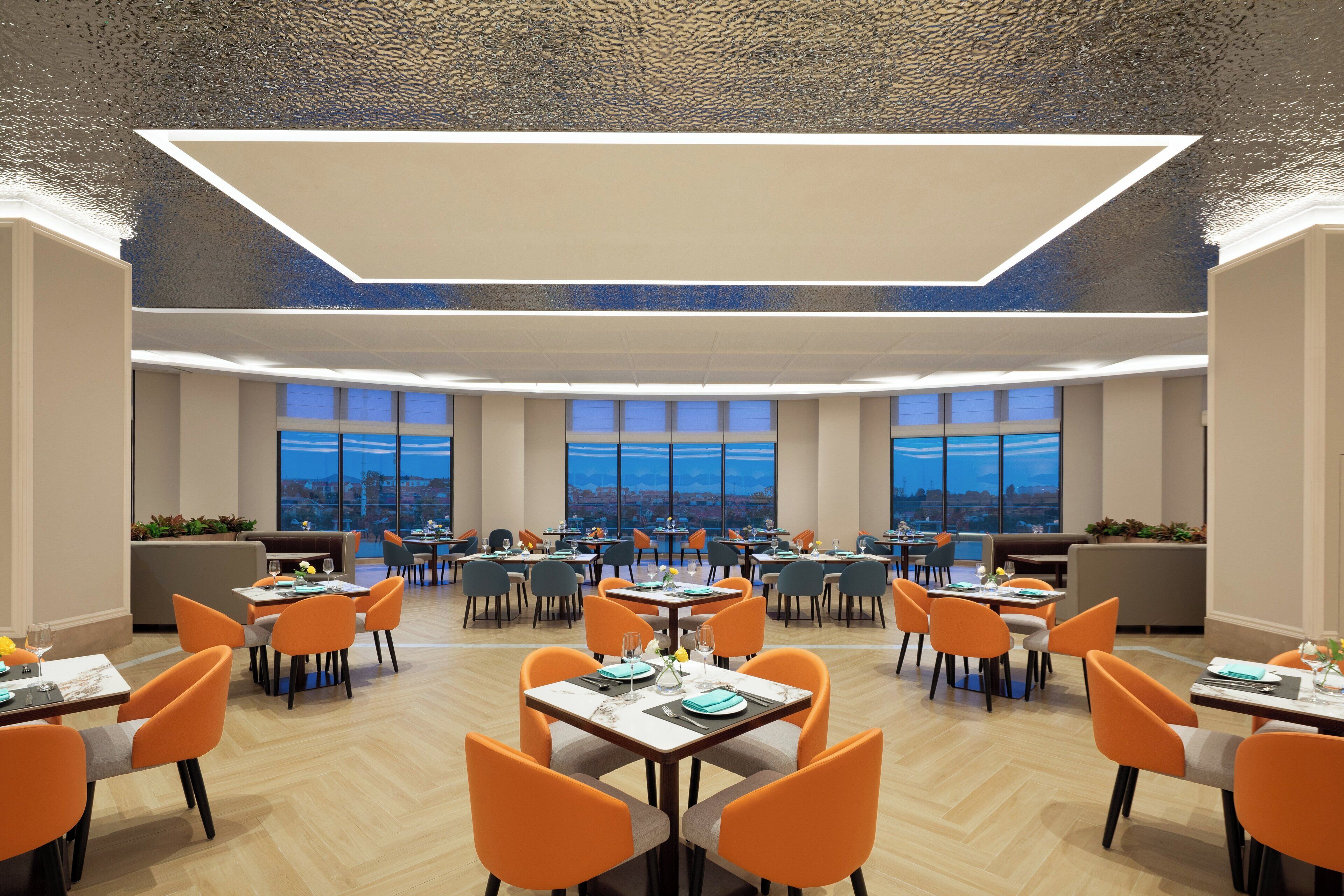 A spacious dining area with multiple tables and chairs, large windows offering a city view, and a modern ceiling design.