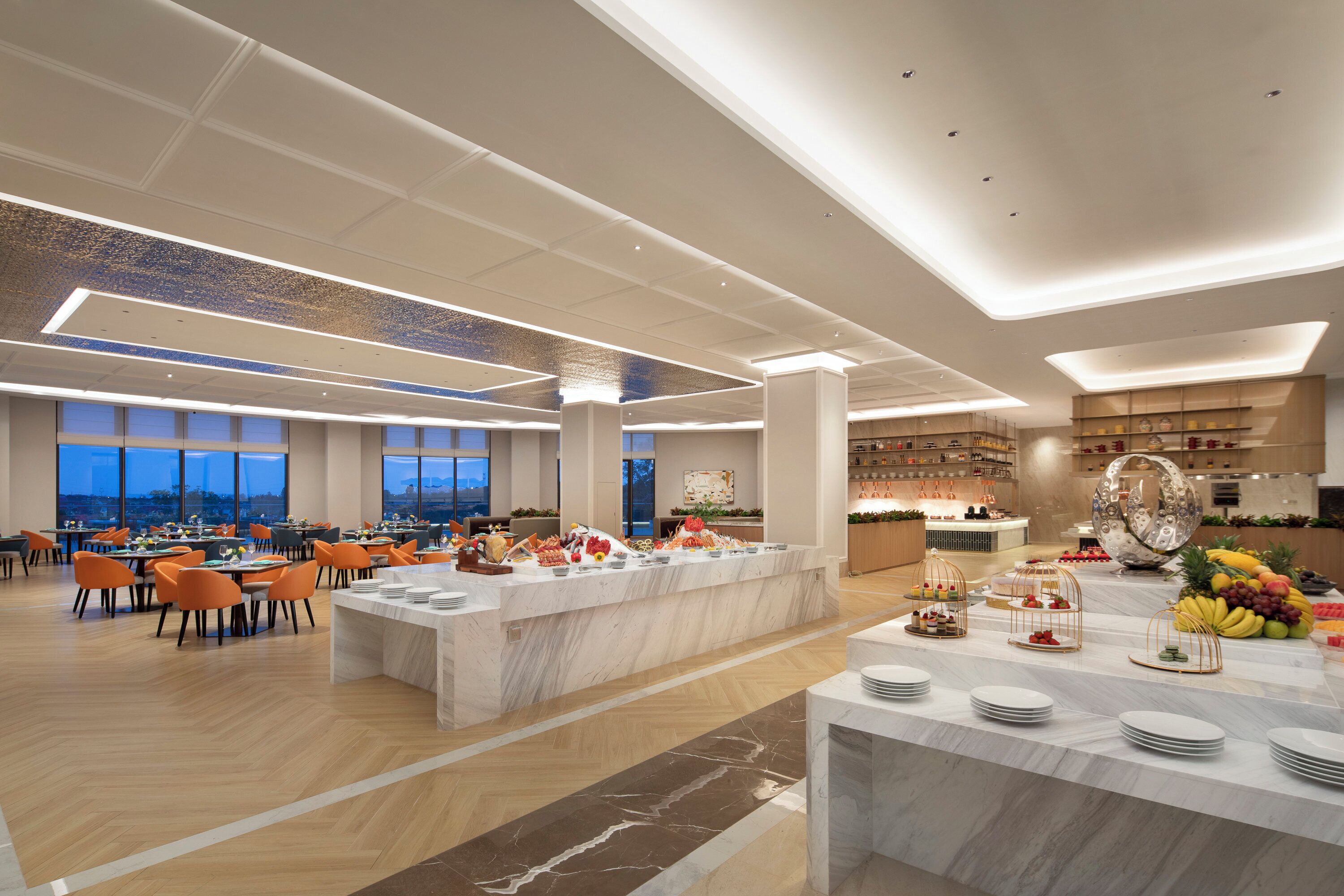 Een ruime eetruimte met meerdere tafels en stoelen, een centraal buffet met diverse gerechten, en uitzicht op de stad door grote ramen.