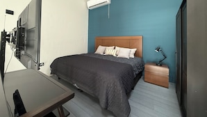 A bedroom with a bed, a desk, a TV mounted on the wall, an air conditioner, and a ceiling fan.