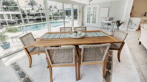Un vaste espace dédié à la salle à manger, avec une grande table en bois et des chaises en osier, donnant sur une piscine et une végétation luxuriante.