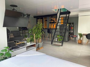 A modern living room with a sofa, a coffee table, a TV, and a ladder. There are several potted plants and a bookshelf in the background.