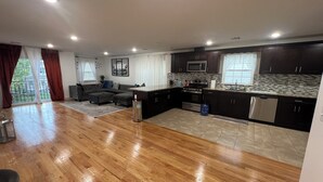 Un vaste espace de vie comprenant un grand canapé d’angle, une cuisine avec des meubles sombres et un plan de travail carrelé, ainsi qu’un plancher en bois.