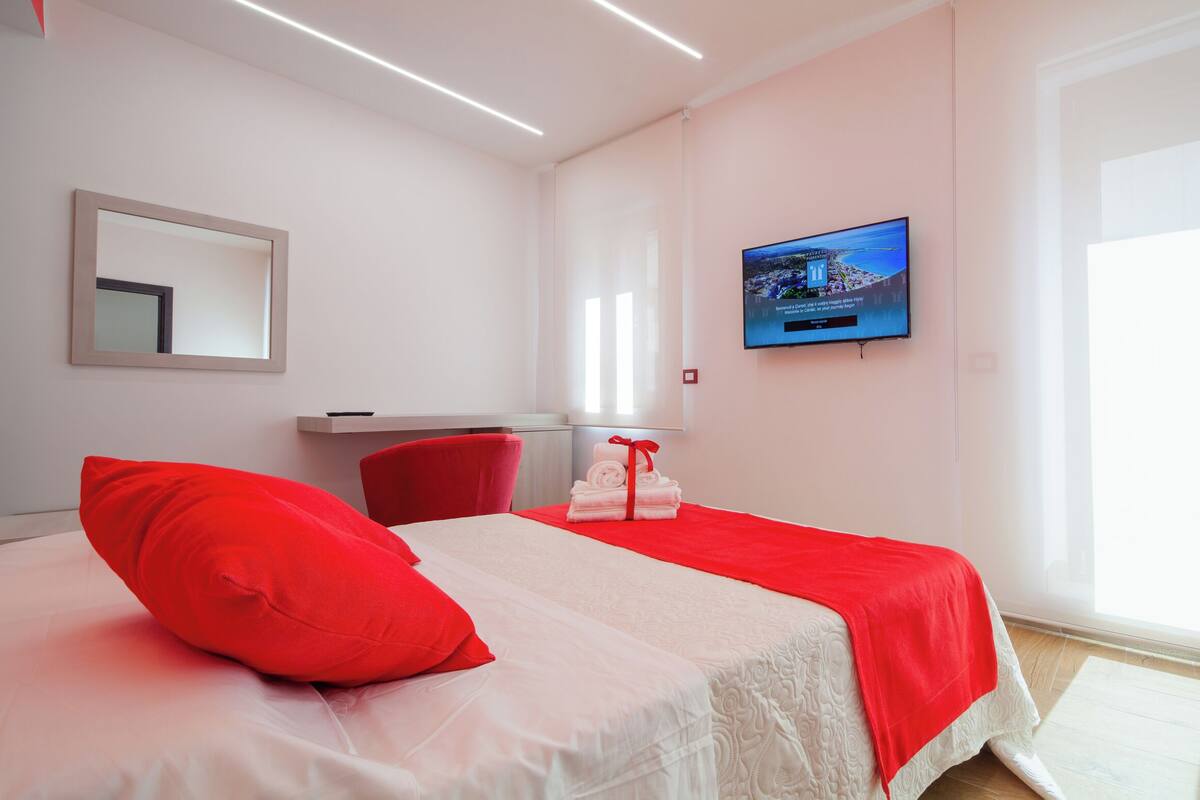 A hotel room with a bed, a red chair, a TV, and a large window.