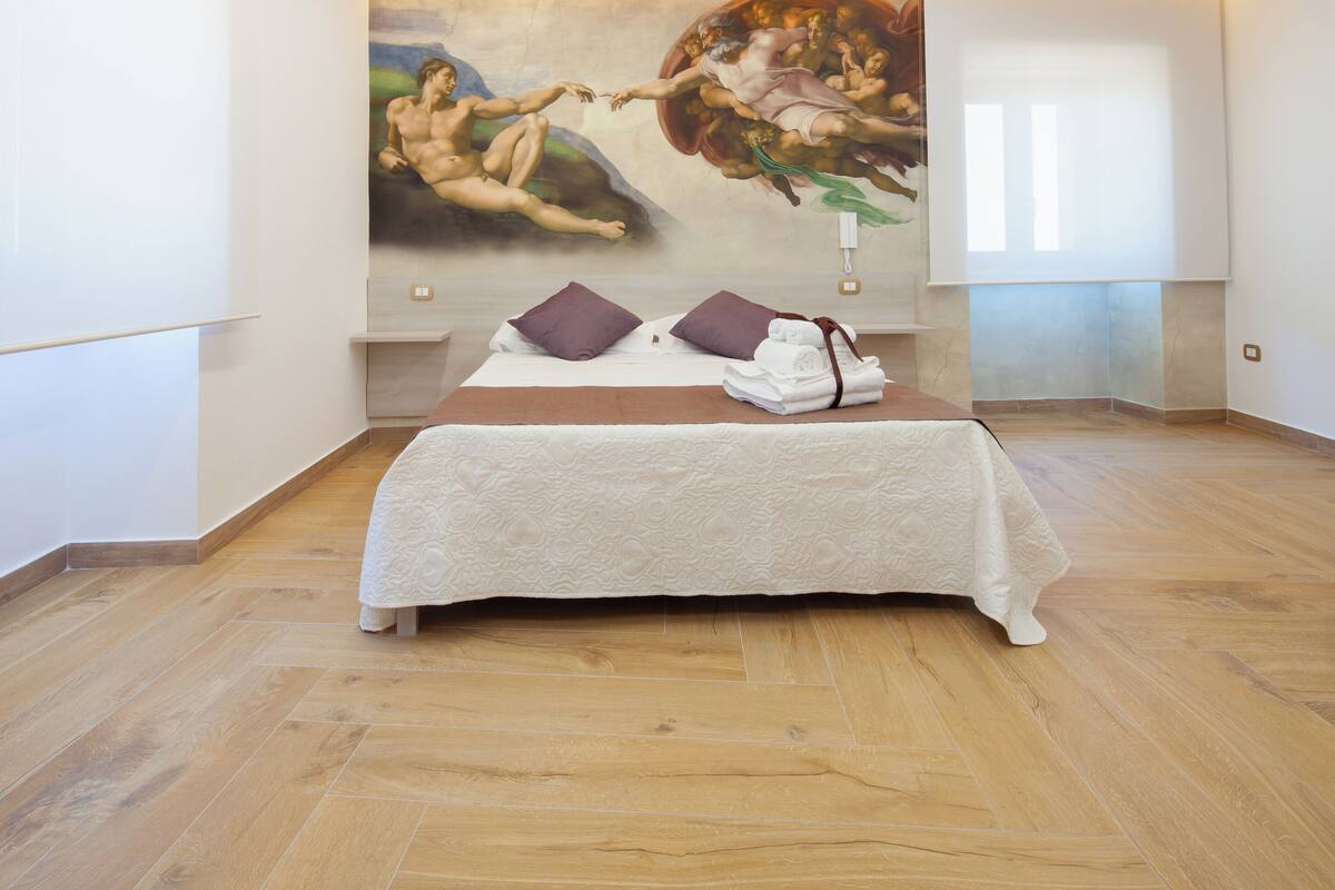 A hotel room with a large mural on the wall, a neatly made bed with pillows, and wooden flooring.