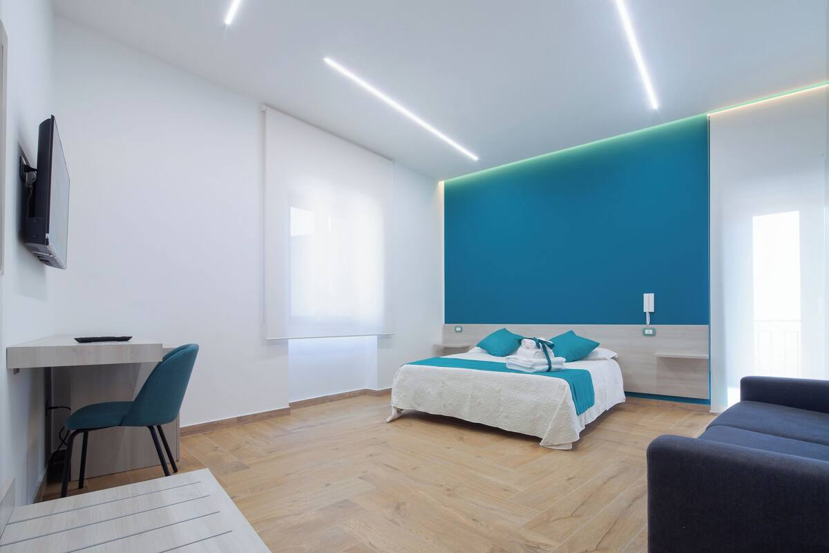 A modern hotel room with a wooden floor, a bed with teal bedding, a desk with a chair, and a TV mounted on the wall.