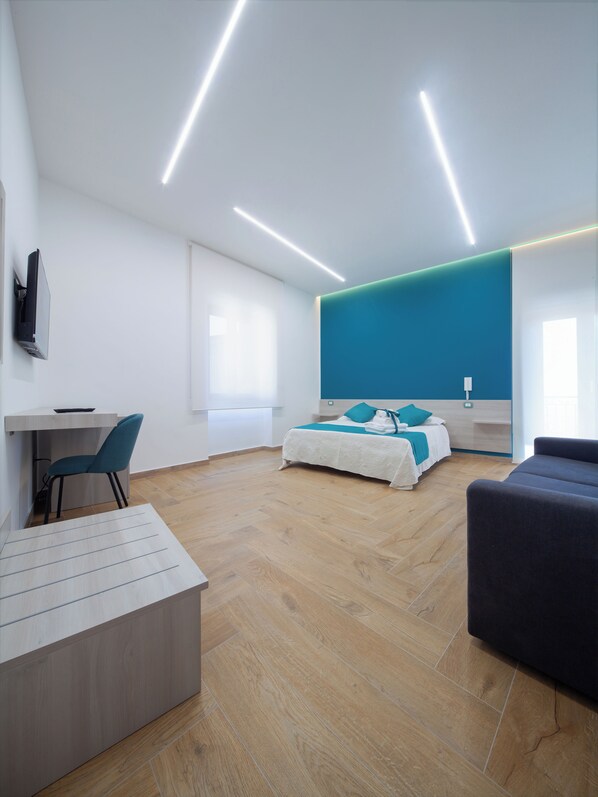 Ein modernes Hotelzimmer mit Holzboden, einem Bett mit türkisfarbenem Bettzeug, einem Schreibtisch mit Stuhl und einem an der Wand montierten Fernseher.