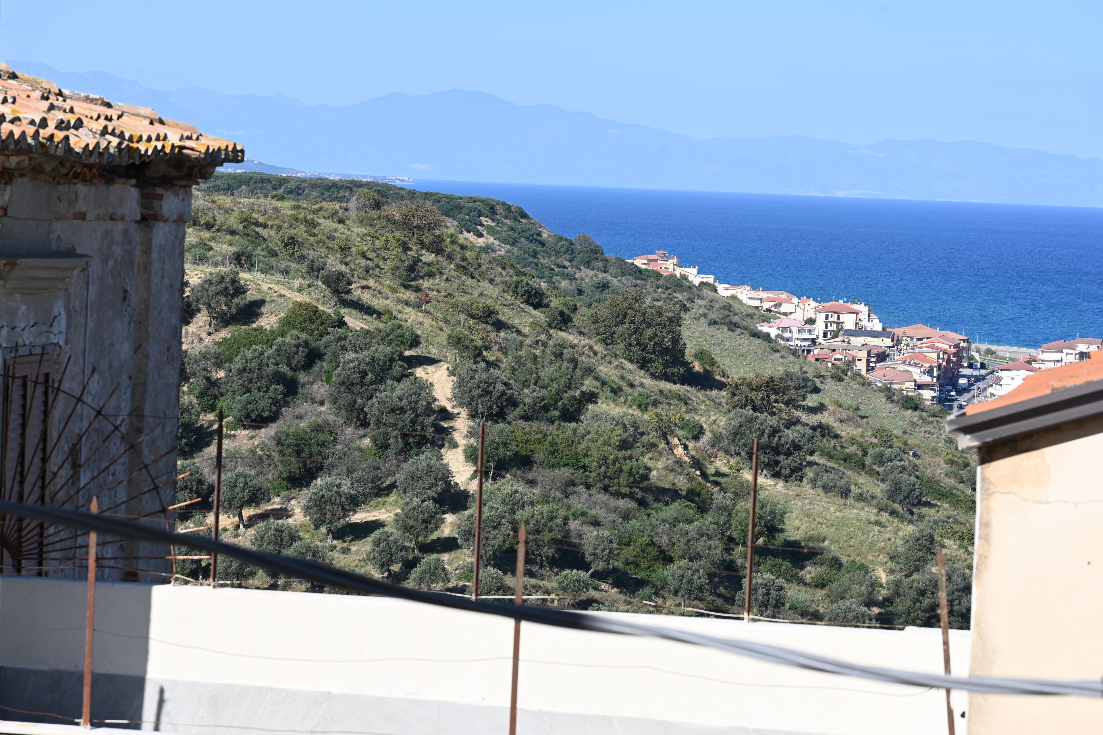 A coastal hillside with buildings, a clear sky, and the sea in the background.
