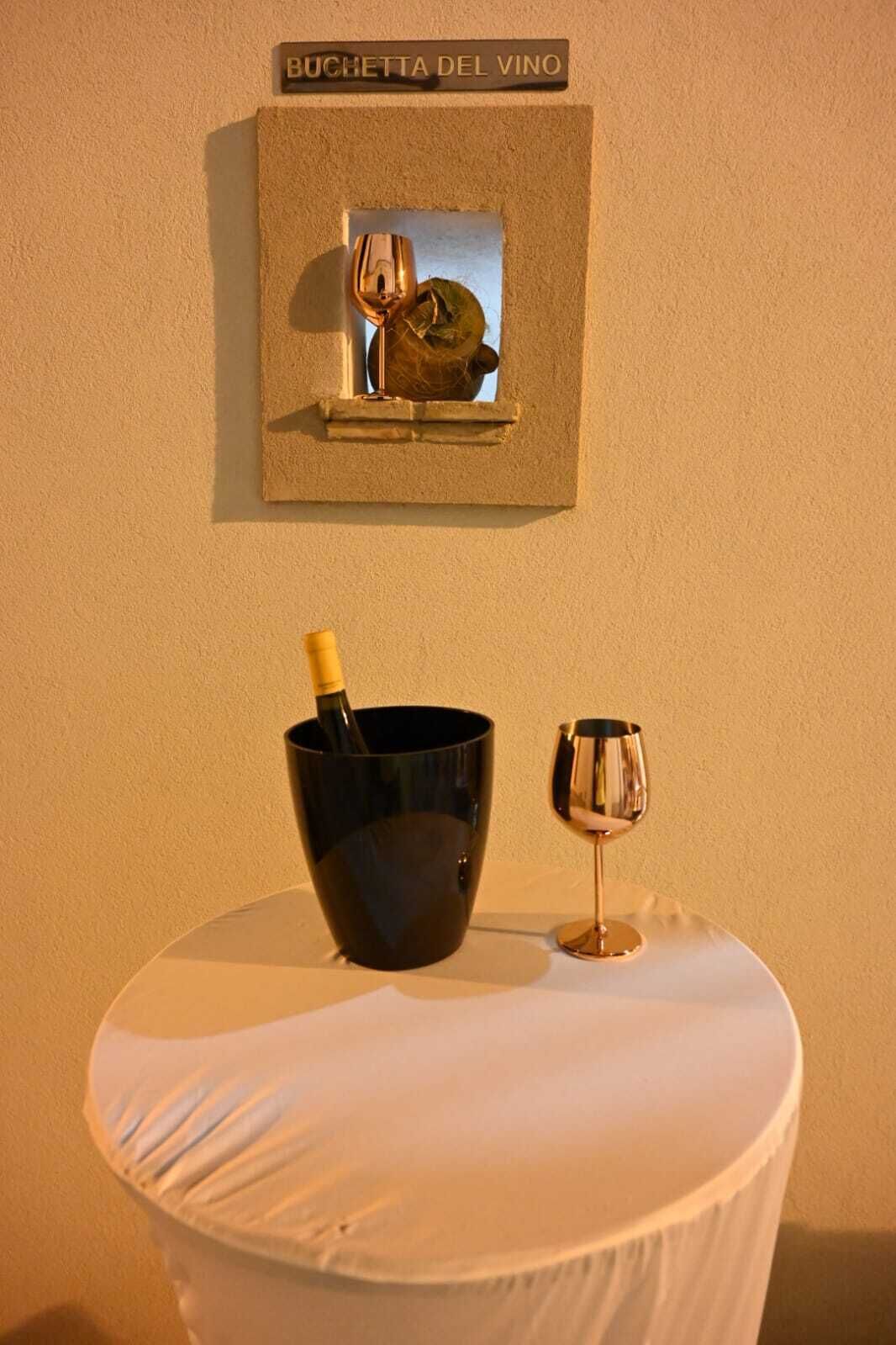 A wall-mounted sign reading "BUCHETTA DEL VINO" above a framed picture of wine glasses, a bottle of wine, and a wine glass on a table with a white tablecloth.