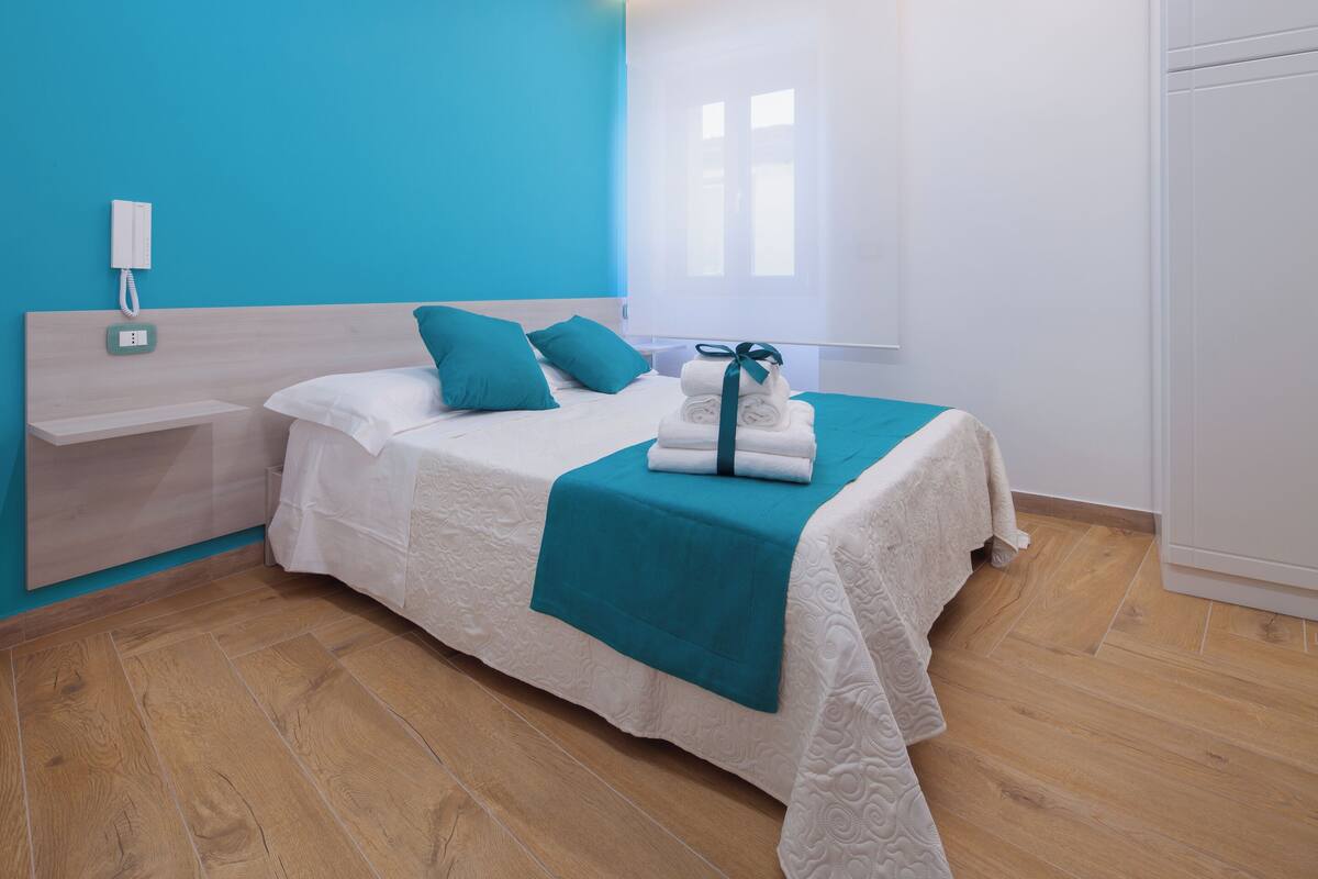 A modern bedroom with a bed, a bedside table, a nightstand, a wardrobe, and a window with blinds.
