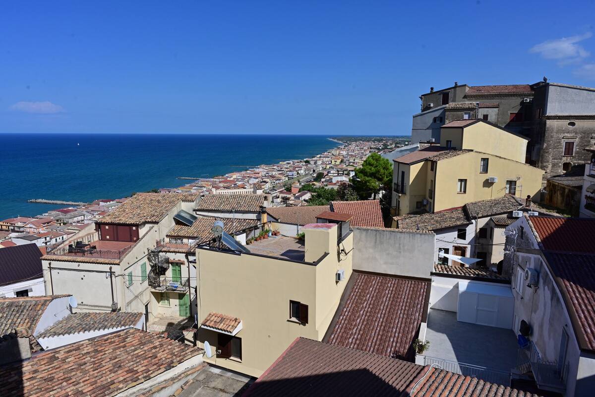 A coastal town with terracotta-roofed buildings, a clear blue sky, and a view of the sea.