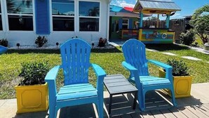 A deck with two blue Adirondack chairs, a small table, and a colorful building with large windows in the background.