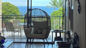 A room with a large sliding glass door leading to a balcony with a view of the ocean. There is a hanging chair and a small table with decorative items. - New updated 2-bedroom condo! Absolutely no stairs! Ocean and golf course views! (Kailua-Kona)
