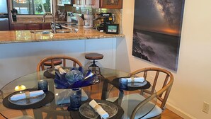 A modern kitchen with wooden cabinets, a granite countertop, and a dining area with a glass table and chairs. - New updated 2-bedroom condo! Absolutely no stairs! Ocean and golf course views! (Kailua-Kona)