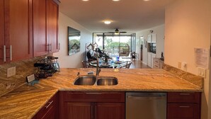 A modern kitchen with wooden cabinets, a double sink, and a dining area with a view. - New updated 2-bedroom condo! Absolutely no stairs! Ocean and golf course views! (Kailua-Kona)