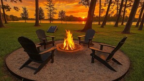 A circular outdoor seating area with a fire pit and Adirondack chairs, surrounded by a gravel border and grass, with a sunset view in the background.