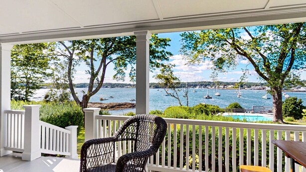 Eine überdachte Veranda mit Blick auf einen See und Boote, ein Korbstuhl und ein Holztisch.